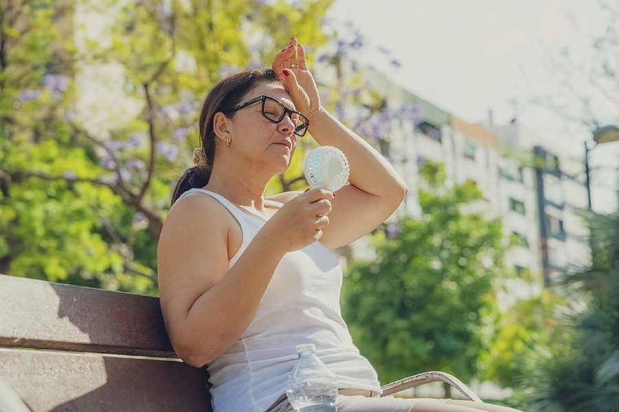 Older woman sits on a park bench in the heat, wiping her forehead while using a small handheld fan.